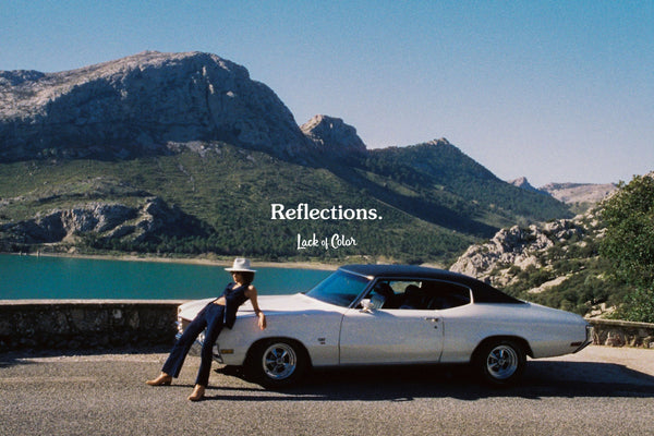 A woman in a hat leans against a white car with mountains and a lake in the background. Text: 'Reflections.' and 'Lack of Color.'