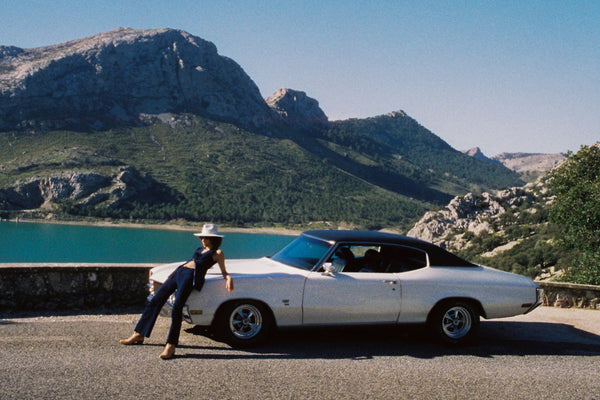 A woman in a white hat leans against a vintage car with mountains and a lake in the background.