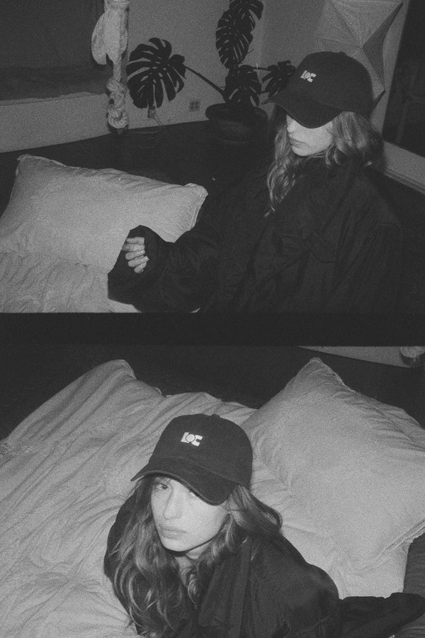 Model wearing the LOC Cap in washed black, sitting on a bed with a relaxed expression.